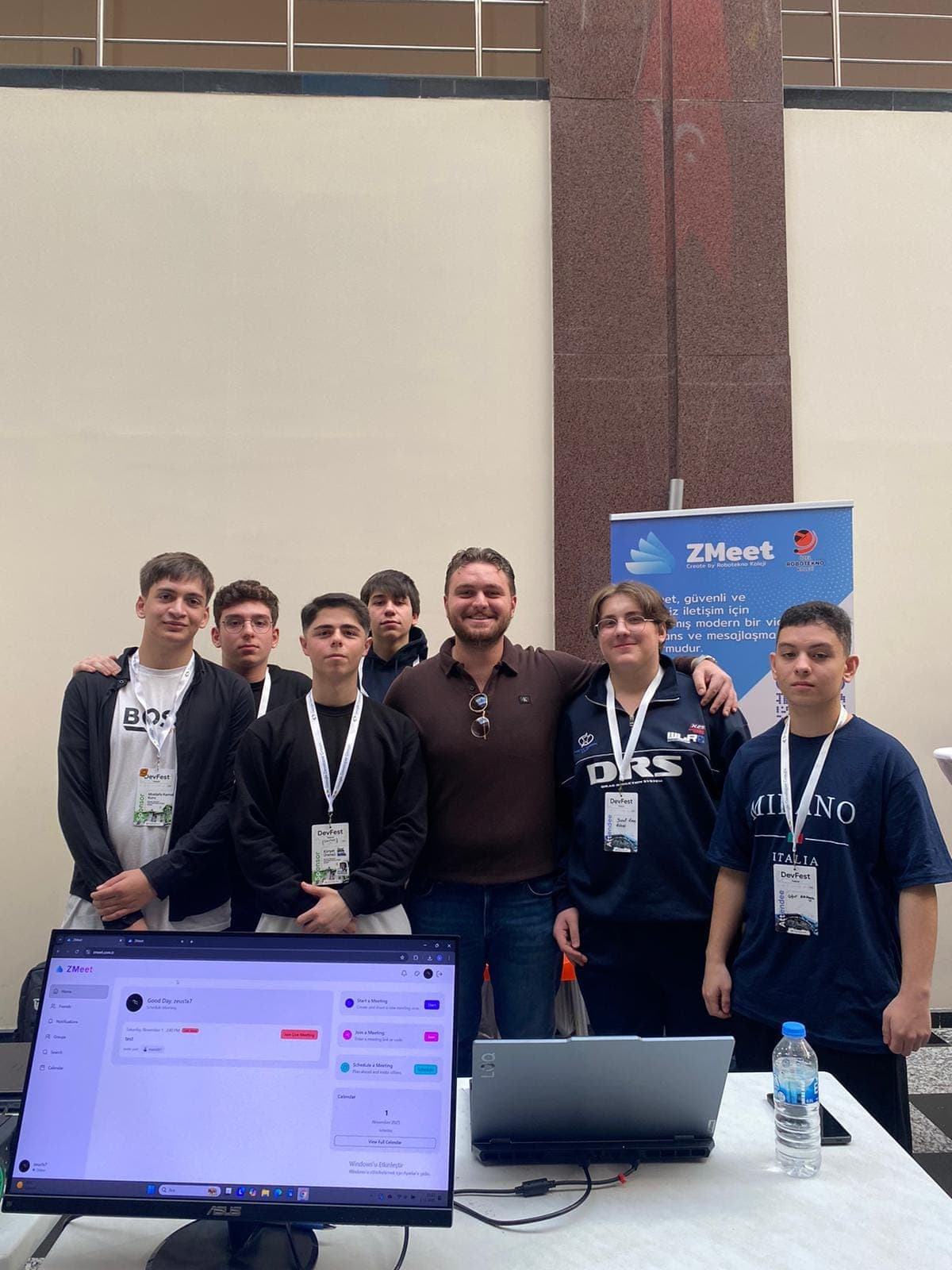 GDG DevFest Trabzon 2025'te ZMeet standının önünde ekip; arkada ZMeet afişi, masada ZMeet arayüzü açık ekran